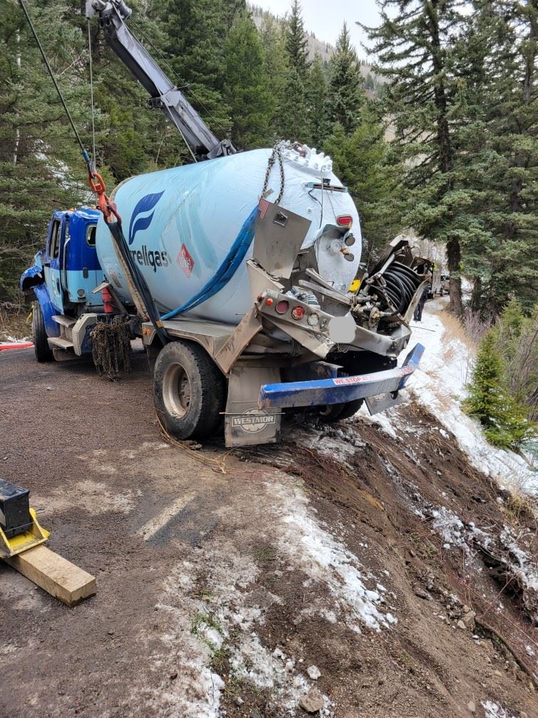 Damaged blue propane truck pulled up from a steep bank by a crane on a tow truck.