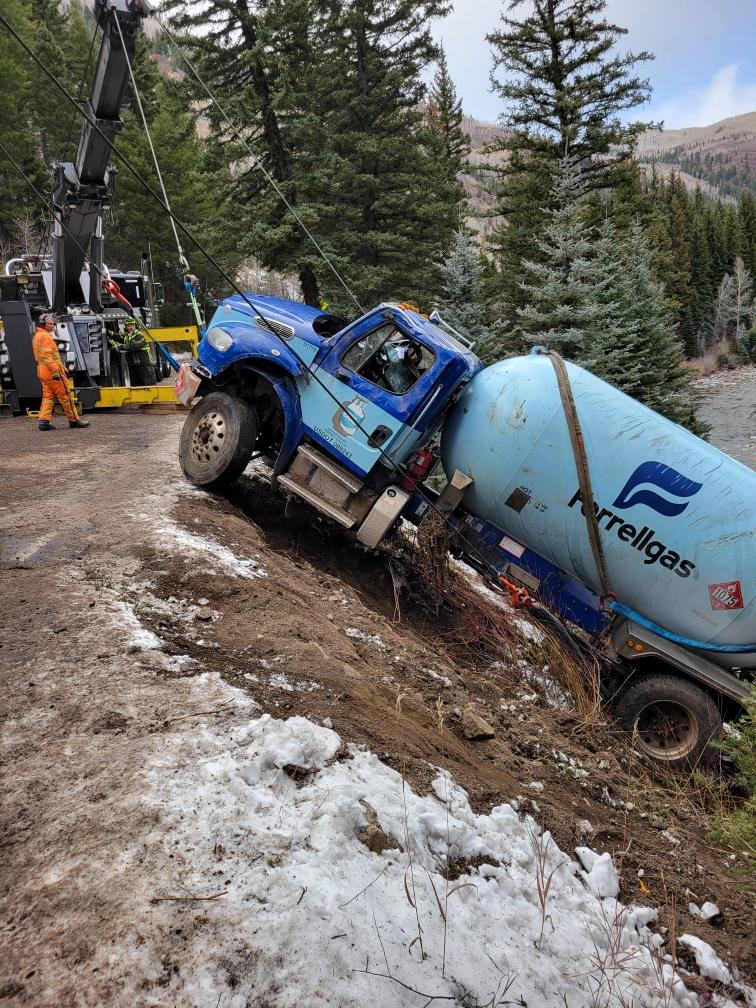 Damaged blue propane truck being pulled up from a steep bank by a crane on a tow truck.